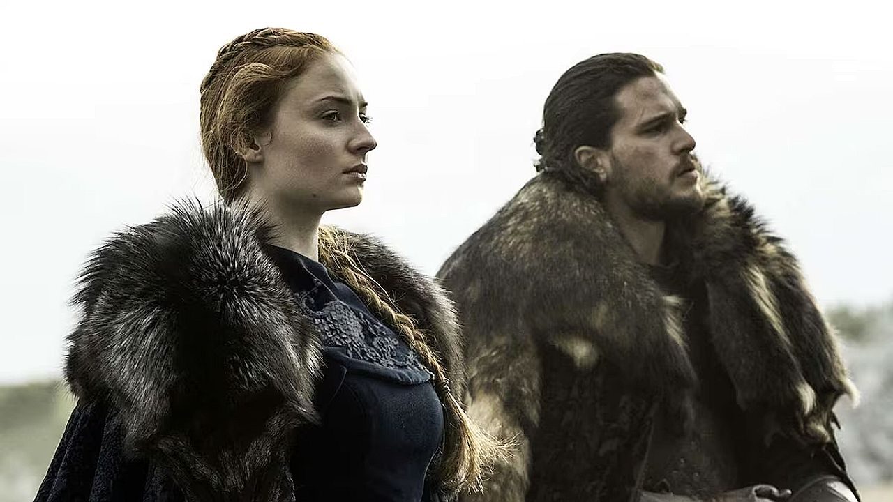 Homem e mulher vestem roupas de época com mantos de pele sobre os ombros, posicionados lado a lado ao ar livre. Ambos olham para o horizonte com expressão séria, em cenário de paisagem fria e céu nublado, sugerindo cena de série ou filme ambientado em contexto medieval ou fantástico.