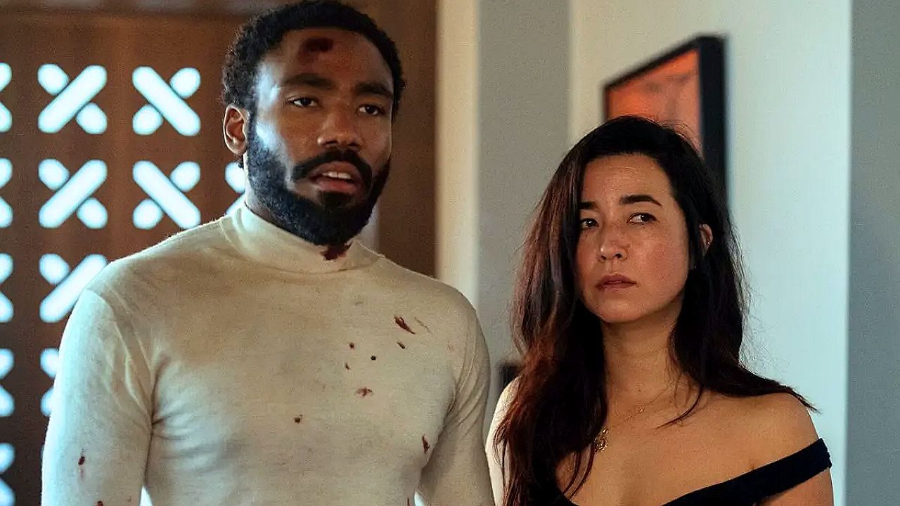 Texto Alternativo disse: Homem com manchas de sangue na roupa e mulher ao lado observam com expressão tensa em cena de série