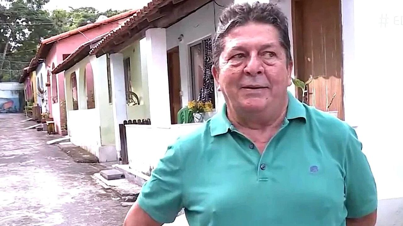 Homem de camisa verde em frente a casas simples fala em reportagem em área residencial