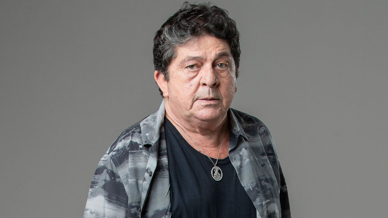 Stepan Nercessian com expressão séria usando camisa estampada aberta sobre camiseta preta em fundo cinza neutro
