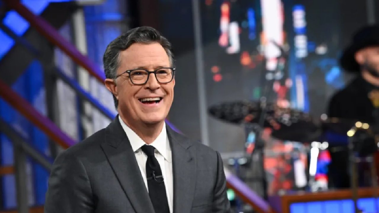 Apresentador de TV veste terno escuro, camisa branca e gravata preta enquanto sorri durante gravação em estúdio. Ele usa óculos de armação fina e está sentado diante de cenário iluminado com tons de azul e vermelho. Ao fundo, é possível ver parte da banda e equipamentos musicais, indicando ambiente de talk show ou programa de entretenimento noturno.