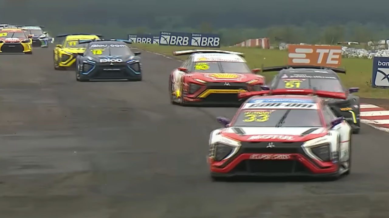 Carros de corrida coloridos da Stock Car em alta velocidade na pista do Autódromo de Campo Grande, com gramado ao fundo e placas publicitárias do BRB