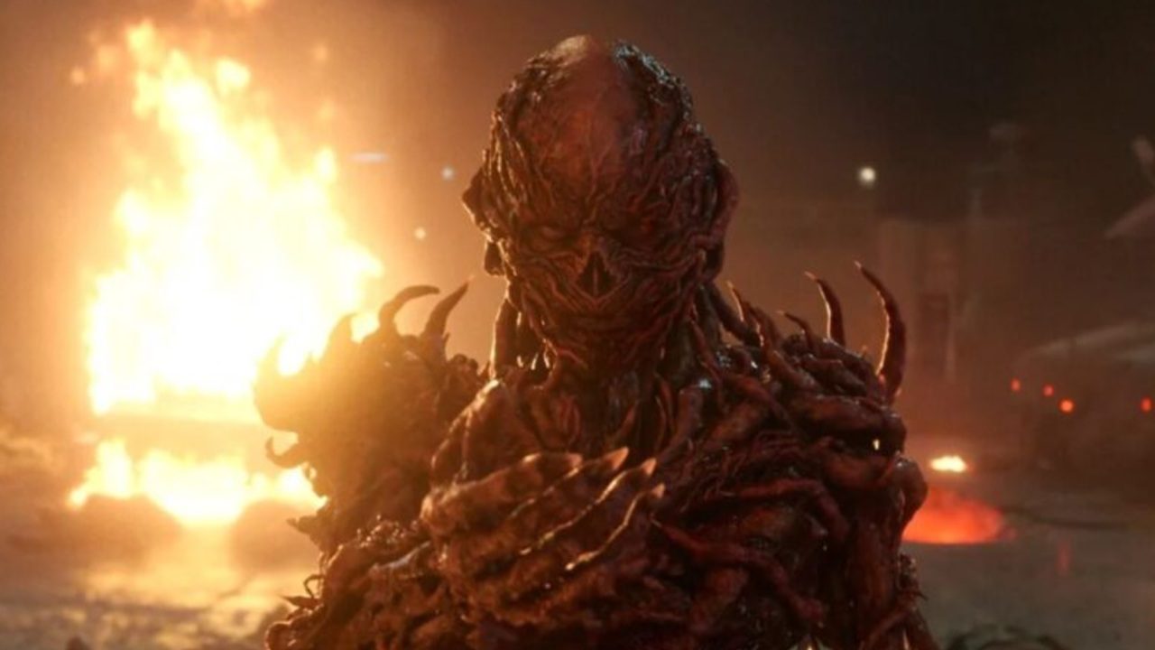 Criatura monstruosa de aparência humanoide, com pele avermelhada e textura orgânica semelhante a raízes retorcidas, aparece em primeiro plano com garras alongadas. Ao fundo, há um incêndio intenso iluminando a cena noturna, criando um clima sombrio e ameaçador