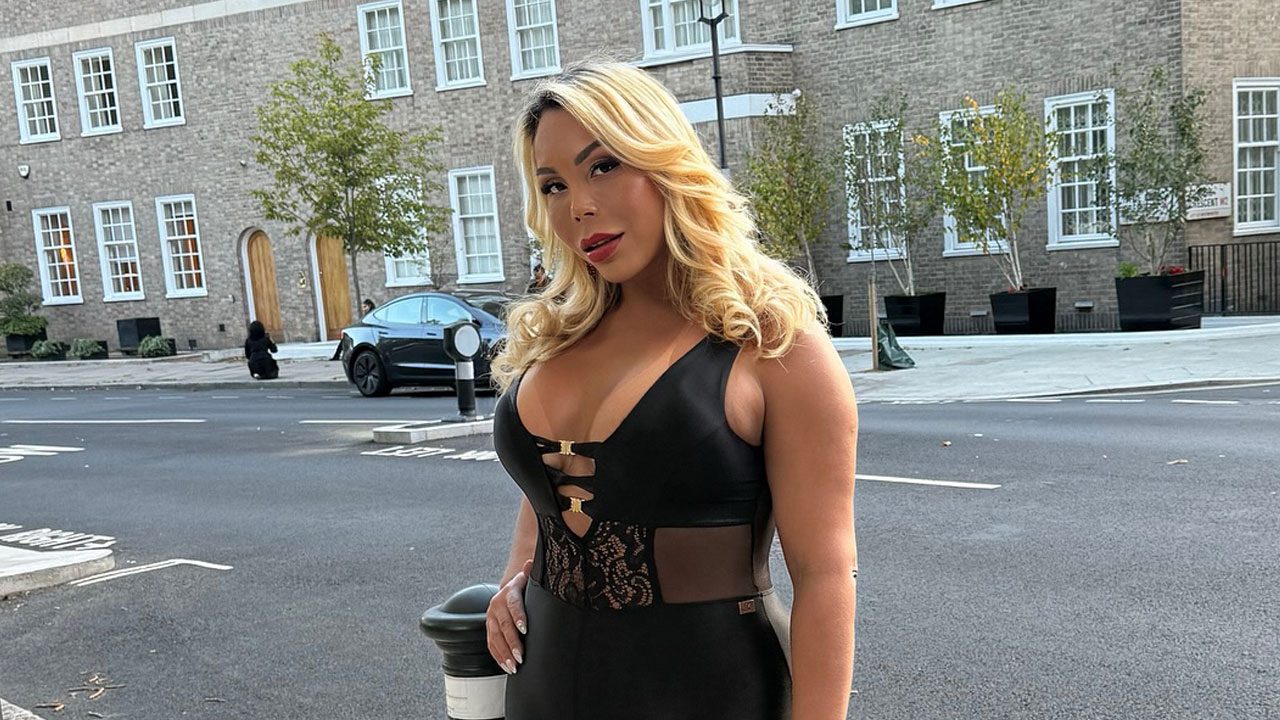 Suellen Carey posando na rua, com cabelo loiro, maquiagem forte e vestindo um body preto de couro e renda