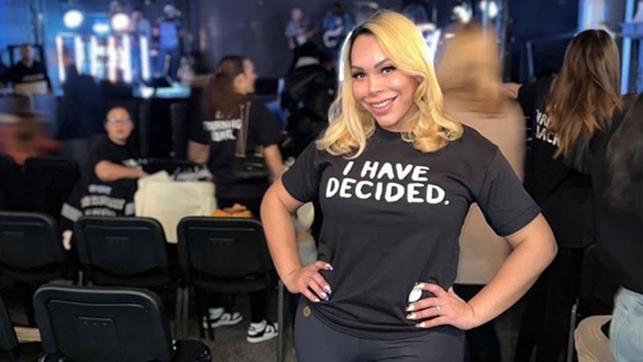 Suellen Carey posa sorrindo com mãos na cintura usando camiseta com frase em inglês durante evento com público ao fundo