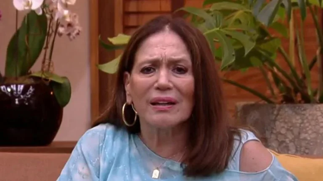 Mulher de meia-idade com cabelos castanhos lisos, expressão preocupada e sobrancelhas franzidas. Ela usa blusa azul clara com ombros à mostra e brincos de argola dourados, sentada em ambiente interno decorado com plantas ao fundo