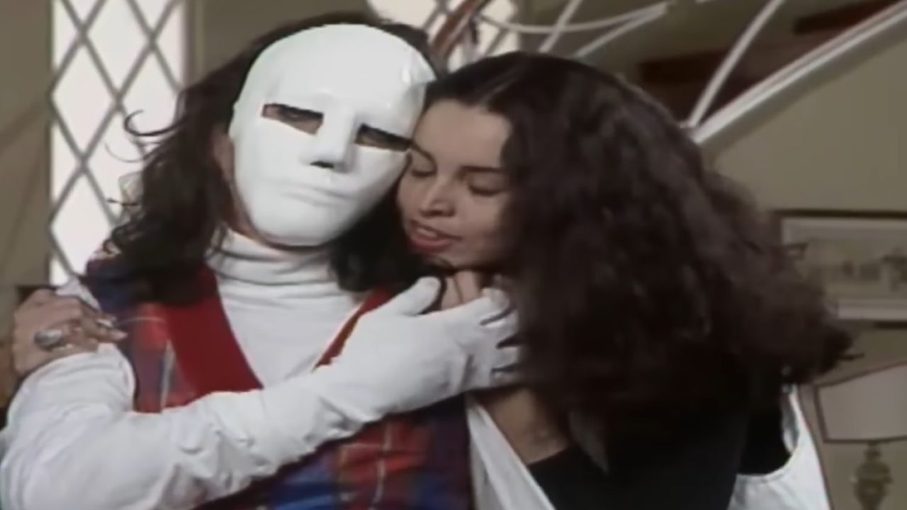 Suzy Rêgo como Carmem em A Viagem ao lado de Mascarado usando máscara branca e figurino com colete xadrez