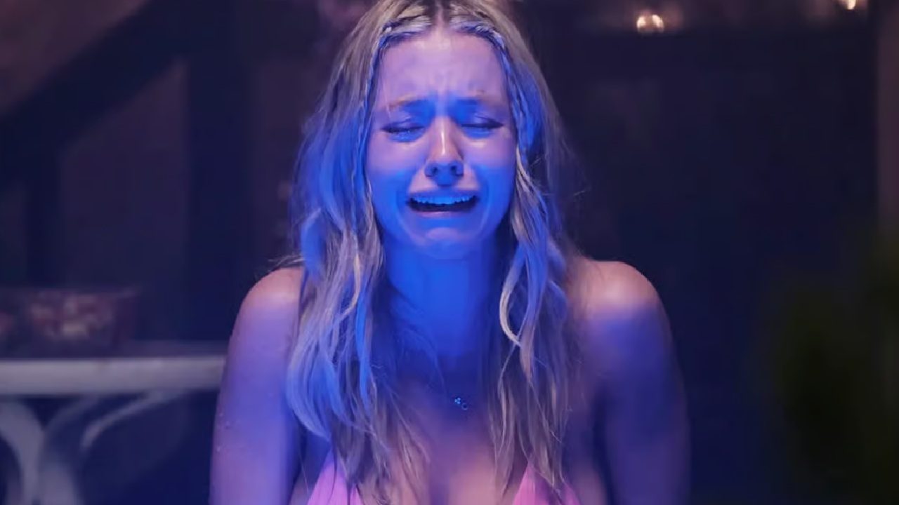 Sydney Sweeney em cena dramática sob luz azul intensa, chorando com expressão de desespero, vestindo roupa rosa em ambiente escuro e iluminado por velas ao fundo