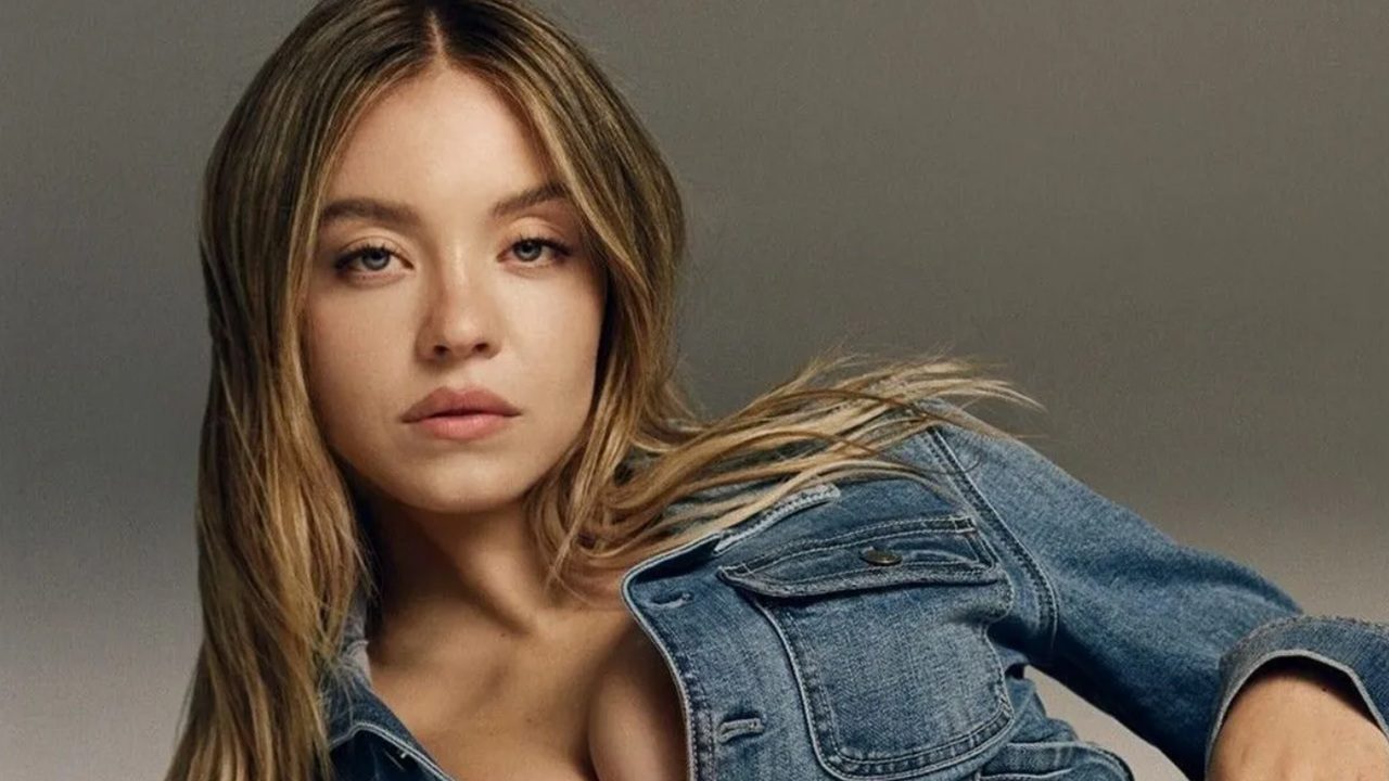 Mulher de cabelos loiros posa com expressão séria vestindo jaqueta jeans diante de fundo neutro