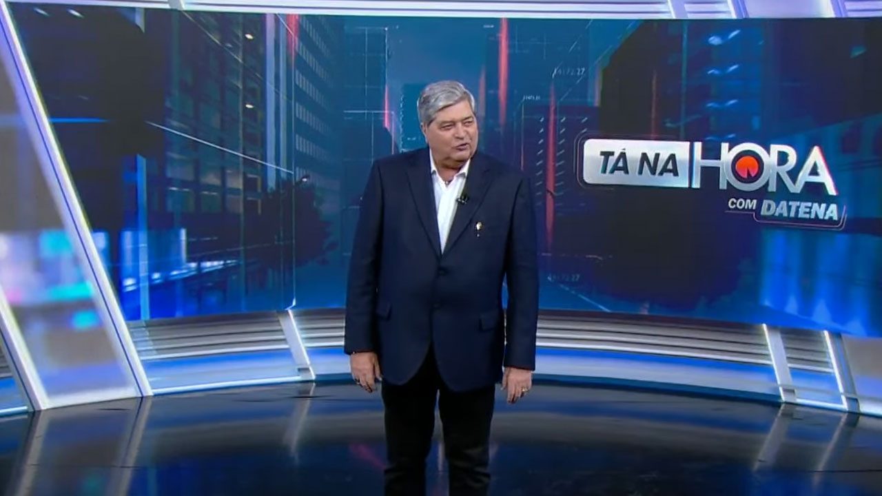 Datena no estúdio do Tá na Hora do SBT em pé diante do telão com o logo do programa ao fundo