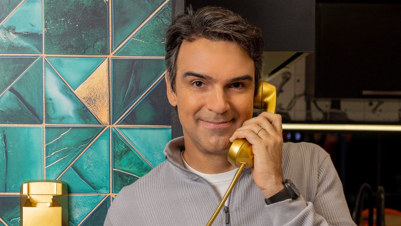 Tadeu Schmidt sorri ao segurar o telefone Big Fone dourado no ouvido, veste blusa cinza em cenário com azulejos geométricos verdes do Big Brother Brasil