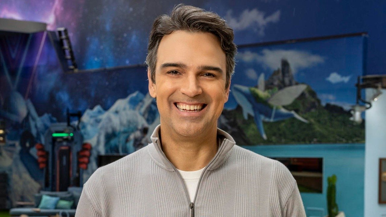 Tadeu Schmidt sorrindo na área externa da casa do Big Brother Brasil com cenário cenográfico colorido ao fundo