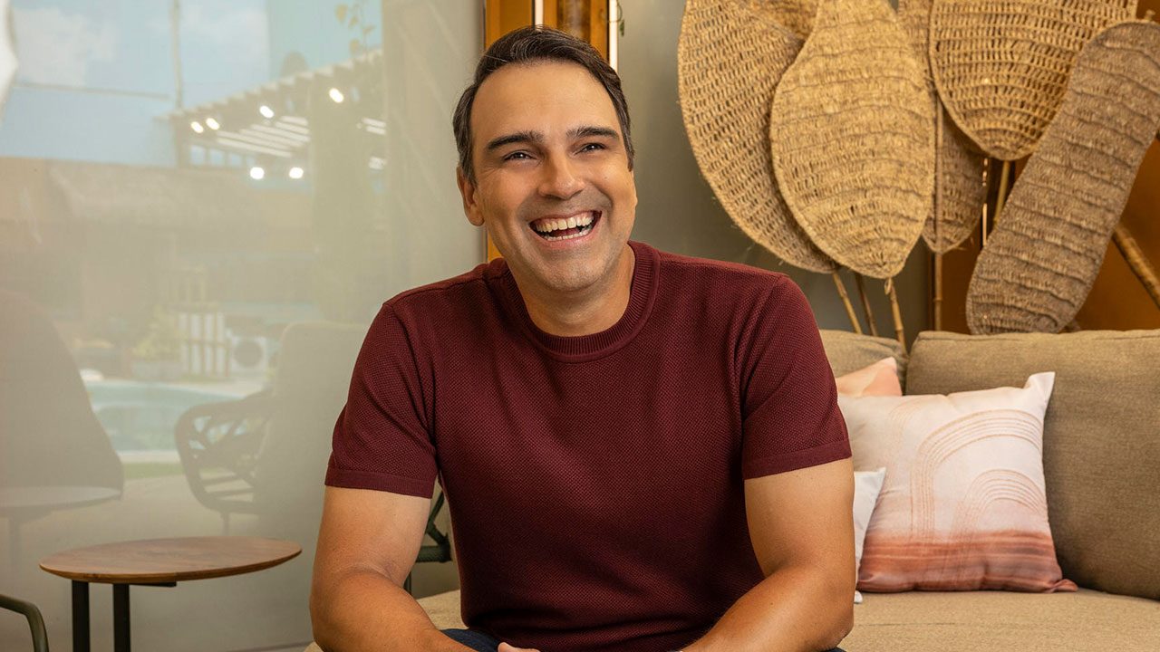 Tadeu Schmidt sorrindo com camiseta vermelha sentado em sofá bege em ambiente decorado com almofadas e fibras naturais