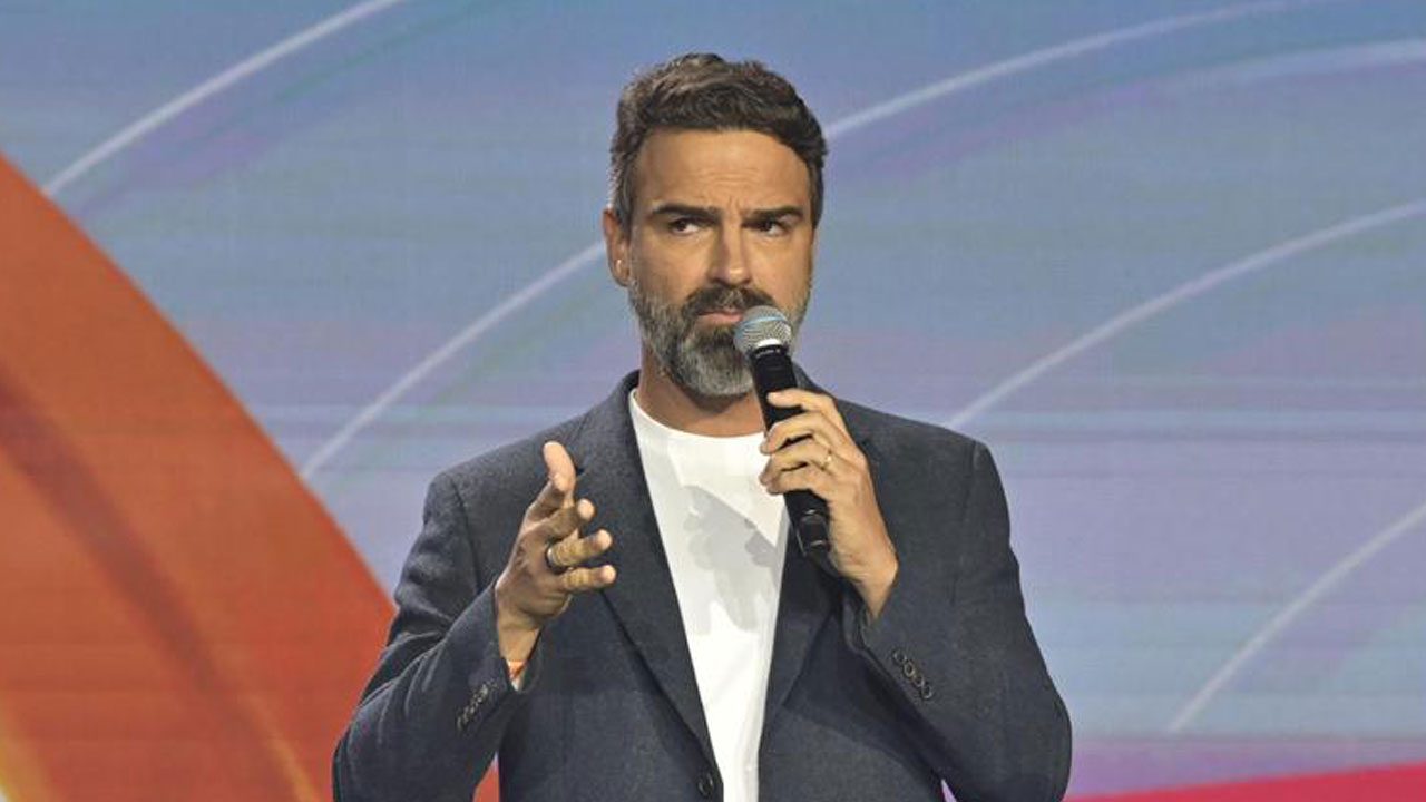 Tadeu Schmidt com barba grisalha e blazer escuro segurando um microfone durante a apresentação de um evento da Globo