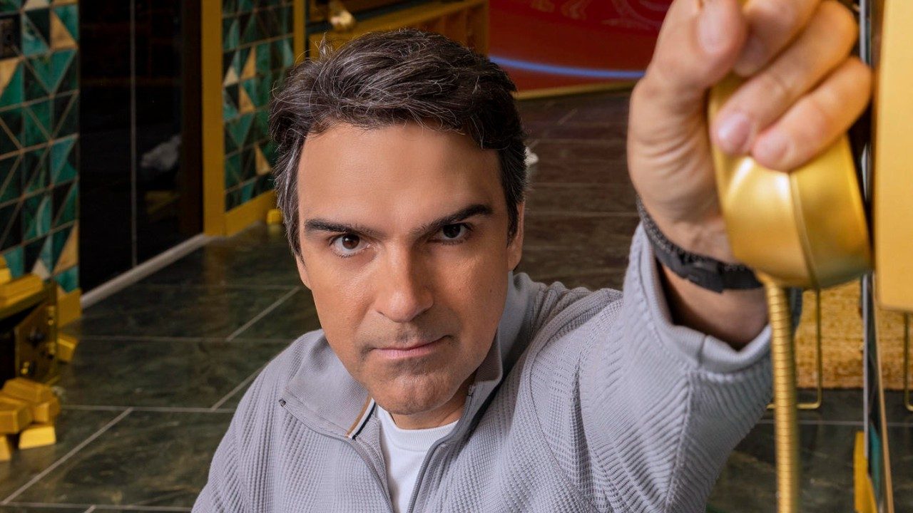 Tadeu Schmidt segura Big Fone dourado com expressão séria em cenário interno com paredes decoradas ao fundo