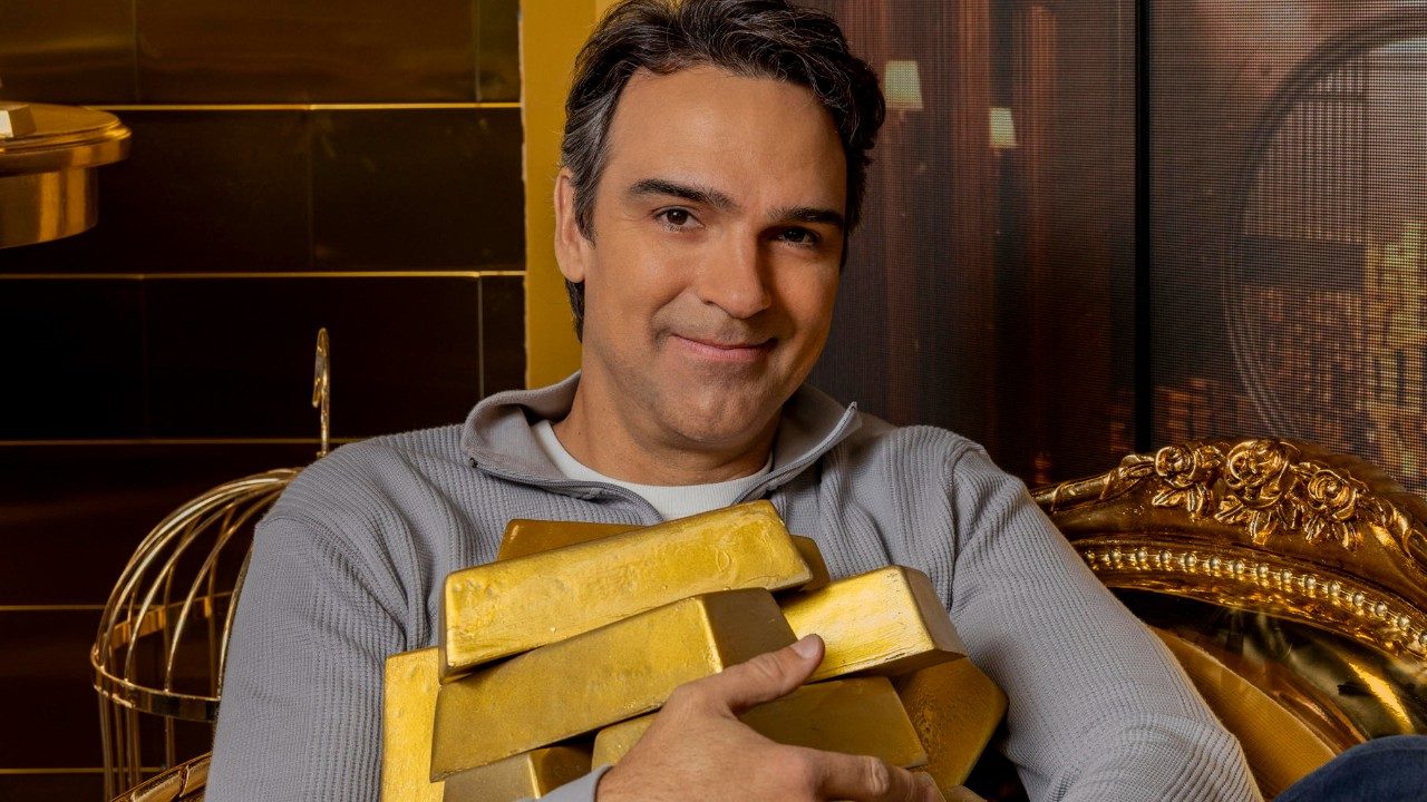 Tadeu Schmidt sorrindo enquanto segura barras de ouro em ambiente luxuoso com decoração dourada ao fundo