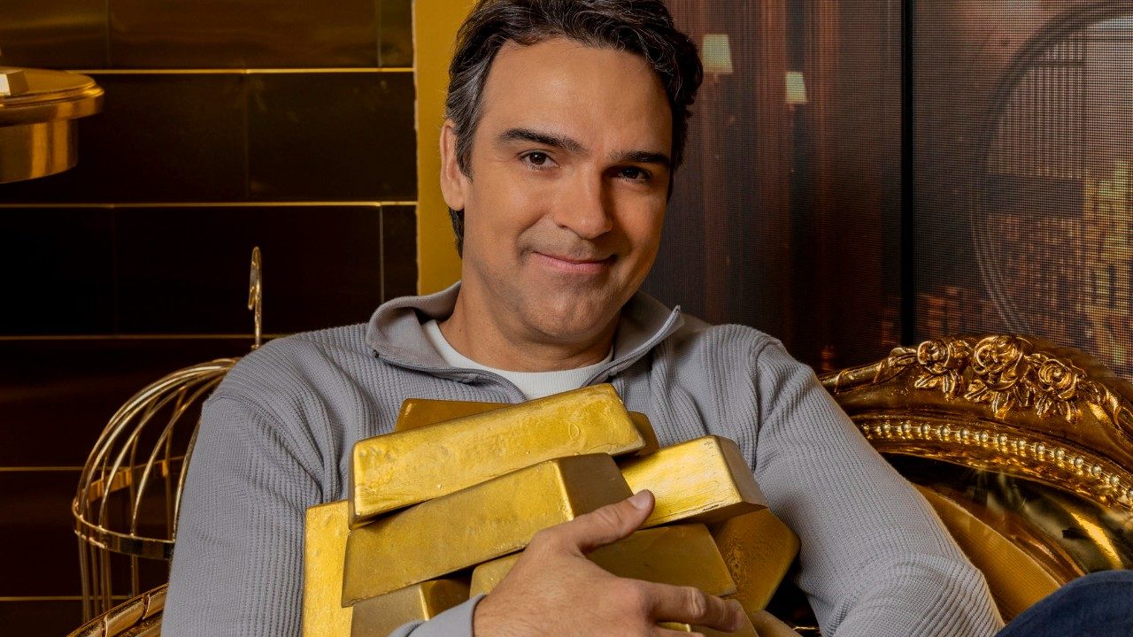Tadeu Schmidt sorri enquanto segura barras de ouro sentado em poltrona dourada