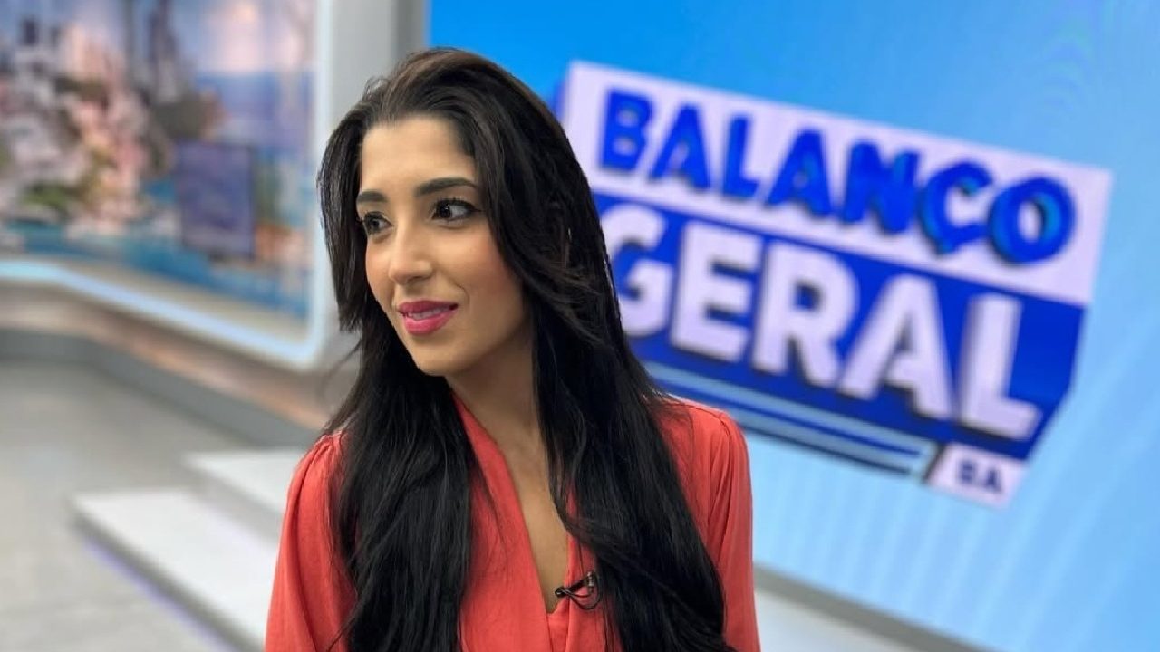 Uma apresentadora de televisão aparece em estúdio, em plano médio, vestindo uma blusa vermelha de mangas longas. Ela olha levemente para o lado, com expressão serena. Ao fundo, um telão azul exibe o logotipo “Balanço Geral”. O cenário é um estúdio de telejornal com iluminação clara e elementos gráficos modernos.