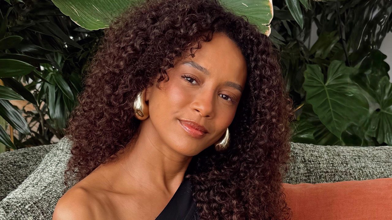 Taís Araujo com cabelo cacheado solto e vestido preto posa sorrindo levemente sentada em sofá com plantas ao fundo