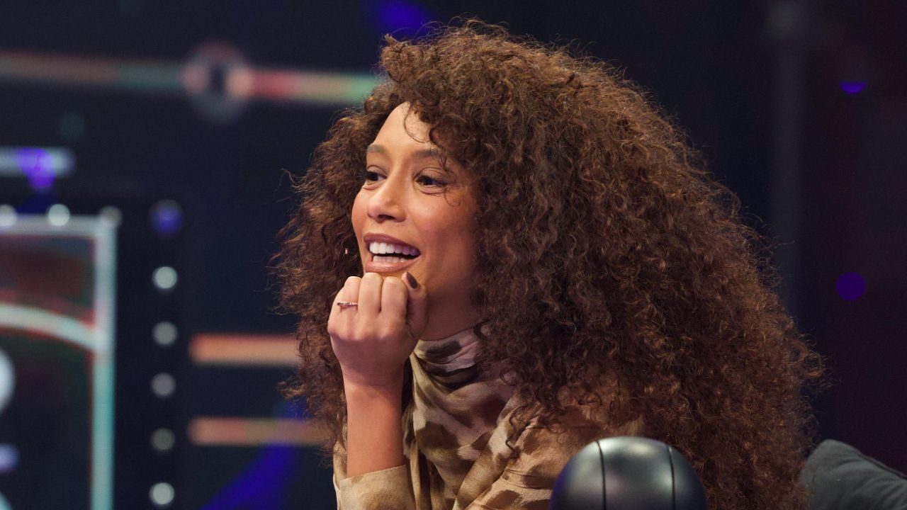 Taís Araujo sorri com a mão no queixo, exibe cabelos cacheados volumosos e veste blusa de estampa animal print em estúdio com luzes desfocadas ao fundo