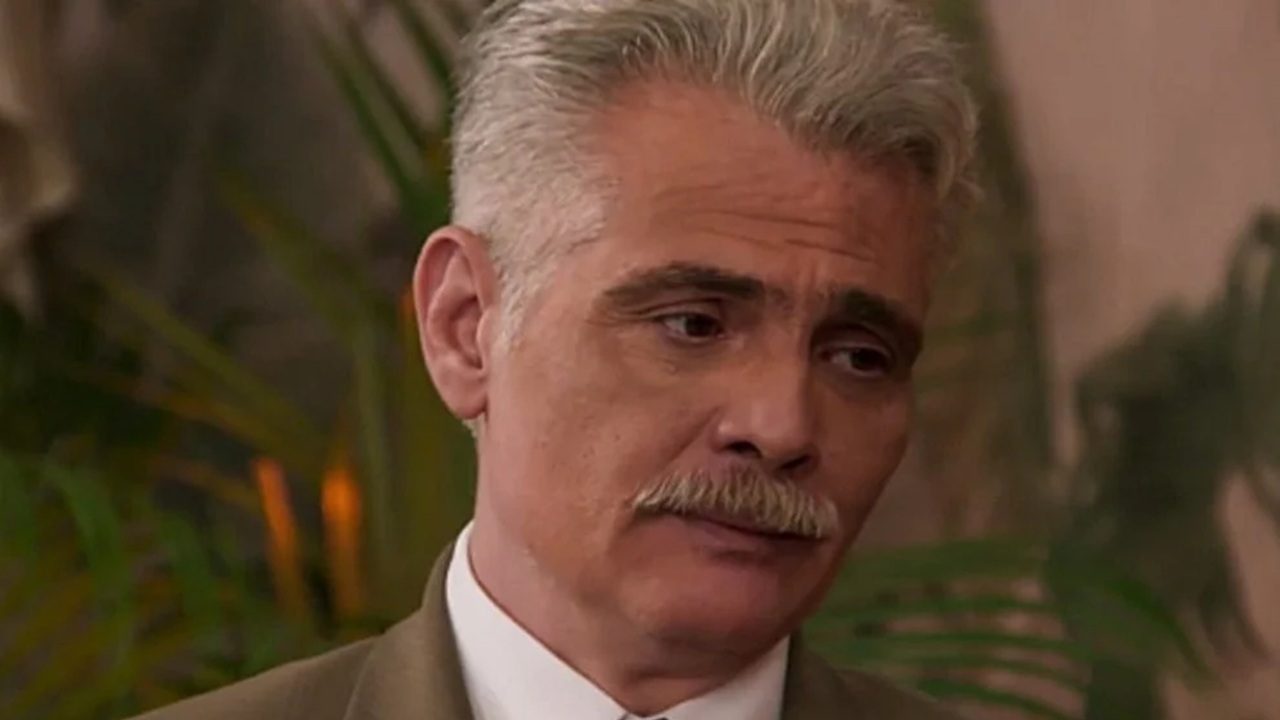 A imagem mostra um homem de cabelo grisalho e bigode, vestindo terno e gravata clara. Ele aparece com expressão séria e olhar levemente pensativo, em um ambiente interno com plantas desfocadas ao fundo, sugerindo uma cena de novela ou série dramática