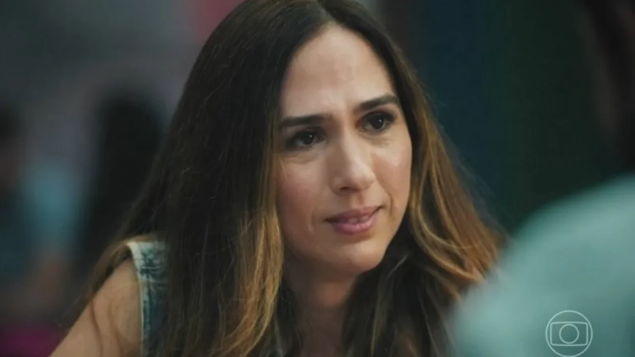 Mulher de cabelos longos e castanhos olha atentamente para alguém fora de quadro, com expressão séria e empática. Ela está em um ambiente interno desfocado, vestindo uma blusa sem mangas em jeans claro. No canto inferior direito, aparece o logotipo de uma emissora de televisão