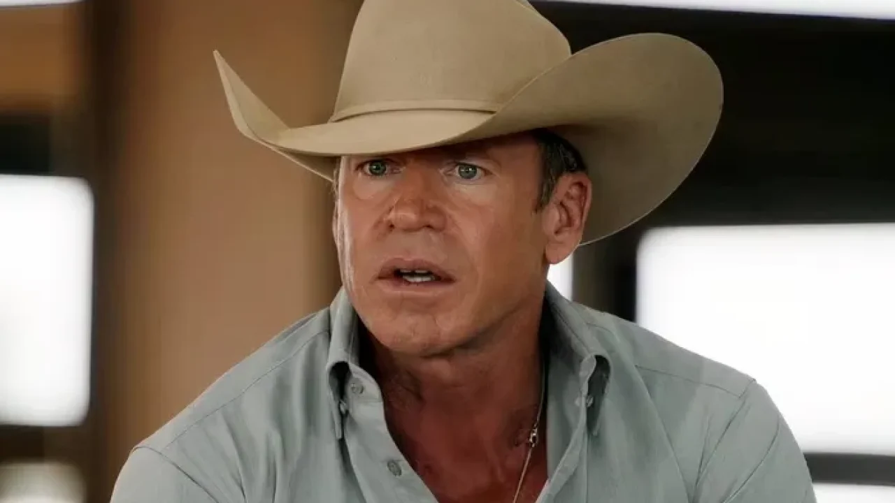 Taylor Sheridan de chapéu de caubói e camisa cinza clara olha com expressão séria, em um ambiente interno bem iluminado