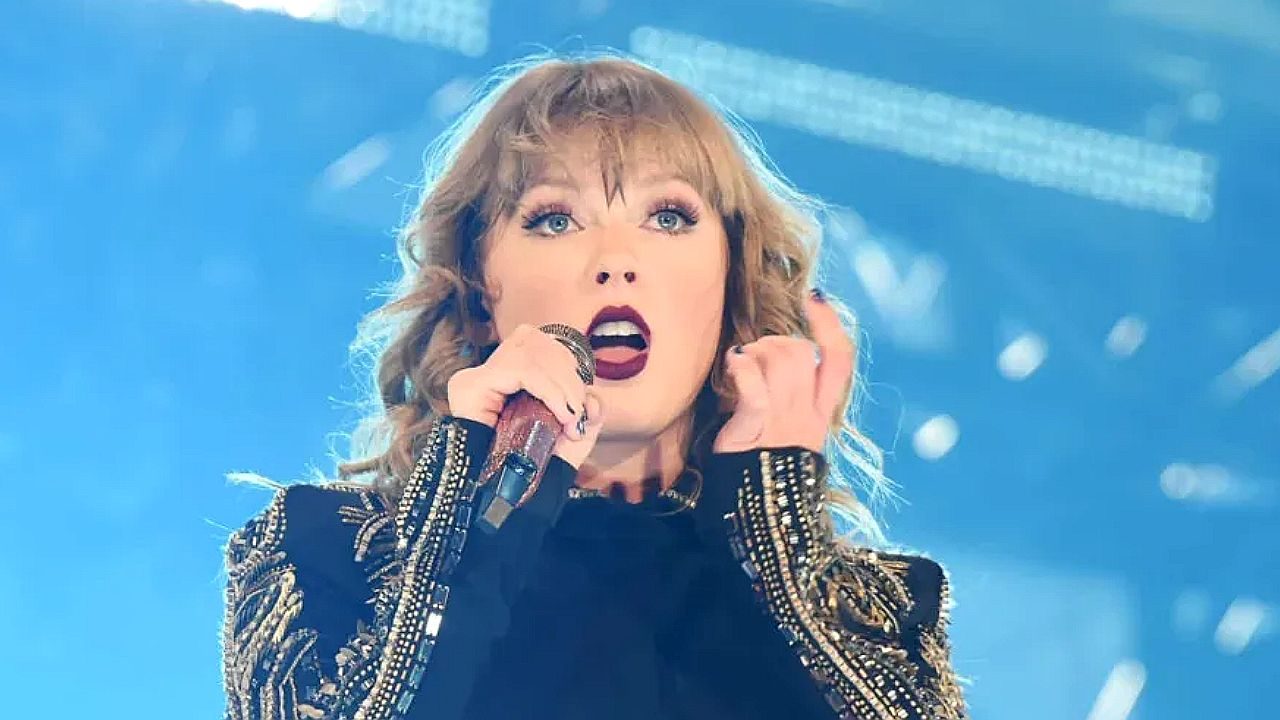 Cantora se apresenta em um show, segurando um microfone com a mão direita e levando a esquerda ao ouvido. Ela usa roupa preta com detalhes brilhantes nas mangas, batom escuro e maquiagem marcante nos olhos. O fundo é iluminado por luzes azuis de palco, criando atmosfera de espetáculo ao vivo.