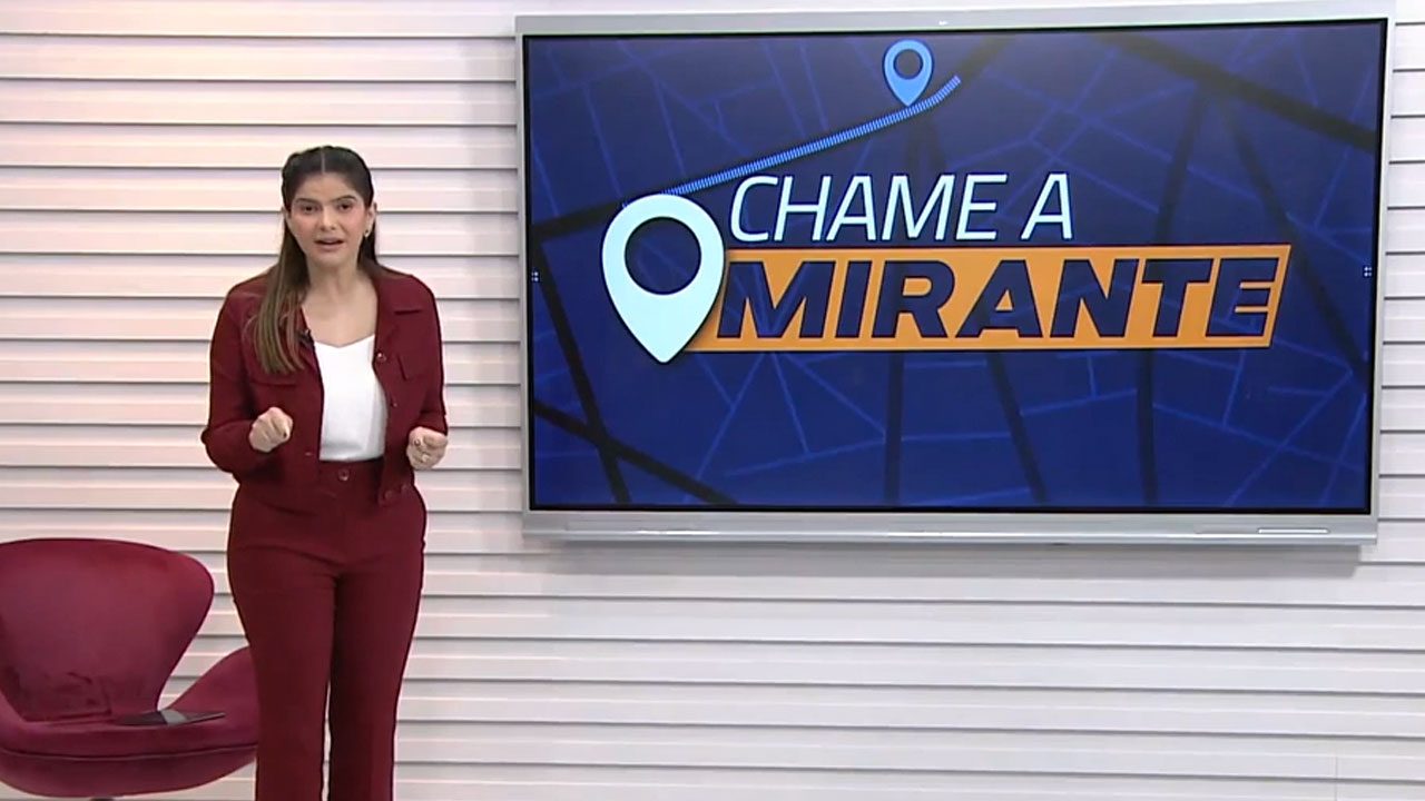 Tayse Feques no estúdio do JM1 da TV Mirante ao lado da tela com o logo do quadro Chame a Mirante