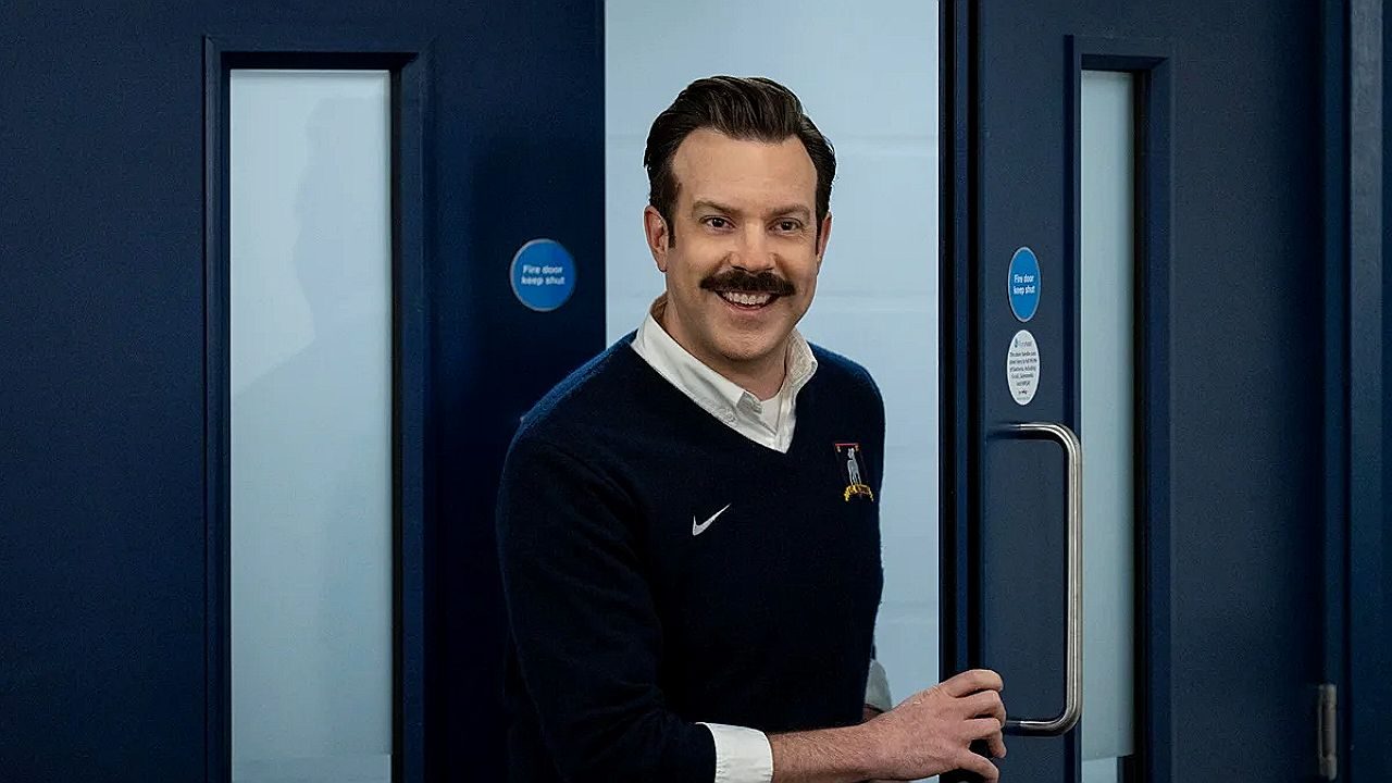 Ted Lasso sorri ao abrir porta do vestiário em cena da série de comédia da Apple TV+
