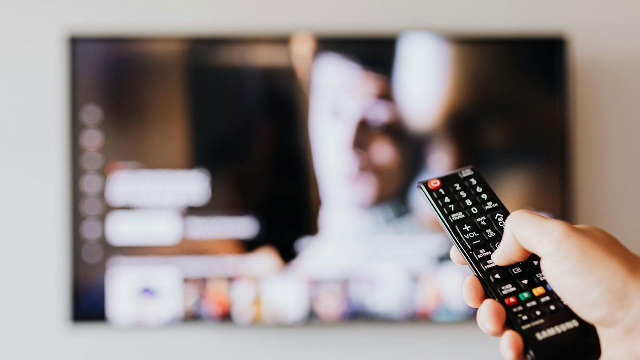 Pessoa segura controle remoto de TV com tela desfocada exibindo plataforma de streaming ao fundo