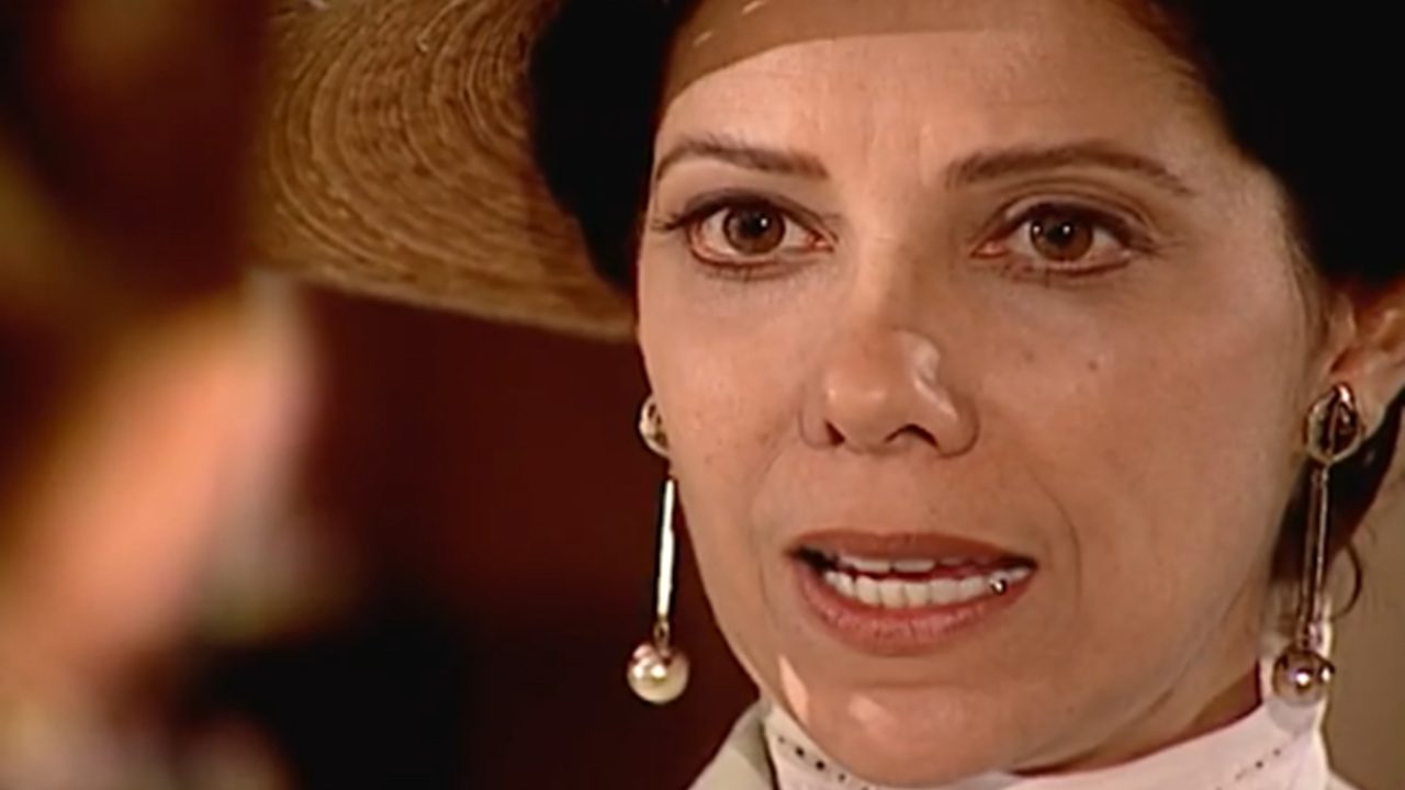 Mulher com chapéu de aba larga aparece em close, olhando com expressão séria. Ela usa brincos longos dourados com pérolas e roupa clara com gola alta. O fundo está desfocado, destacando o foco em seu rosto