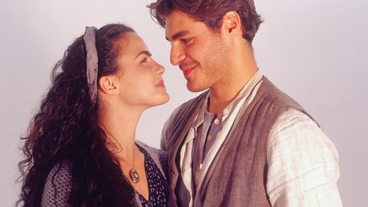 Homem e mulher se encaram sorrindo de forma carinhosa, em clima romântico, vestindo roupas simples de tons neutros e iluminação suave ao fundo