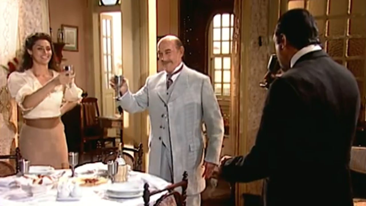 Três pessoas brindam sorrindo em torno de uma mesa posta em sala iluminada; o homem ao centro veste terno claro, enquanto os outros dois, uma mulher e um homem, seguram taças em clima de celebração