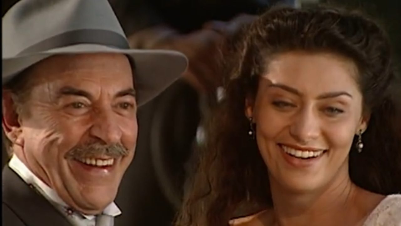 Homem mais velho, usando chapéu cinza e terno, sorri amplamente ao lado de uma mulher jovem de cabelos longos e escuros, também sorridente. Ambos aparecem em um ambiente interno com luz quente, transmitindo clima de alegria e cumplicidade