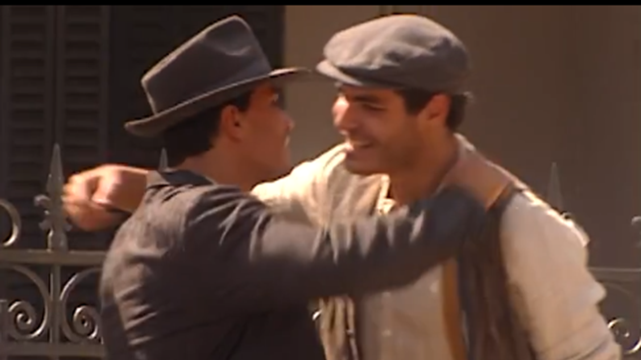 Dois homens se cumprimentam com um abraço caloroso em ambiente externo ensolarado, ambos usando chapéus e roupas de época, sugerindo uma cena de reencontro ou amizade