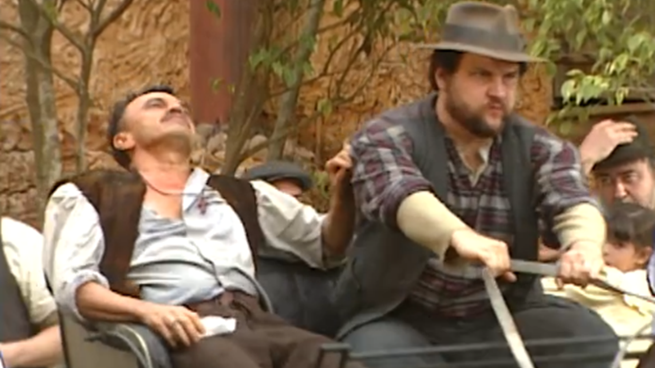 Cena de época mostra um homem desacordado sendo transportado em uma carroça conduzida por outro homem, com expressão tensa, enquanto pessoas observam ao fundo