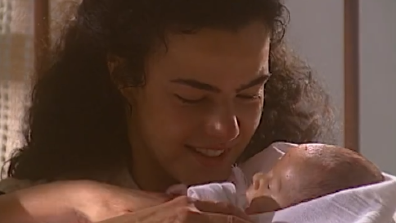 Mulher sorri emocionada enquanto segura um bebê recém-nascido enrolado em um pano branco, em cena iluminada suavemente que transmite ternura e afeto