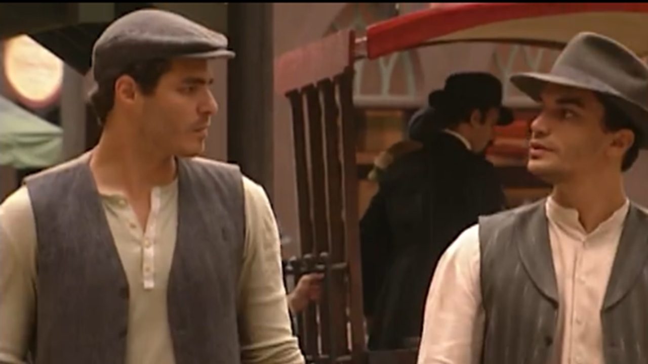 Dois homens caminham lado a lado em uma rua de época, usando roupas antigas — camisas claras, coletes e boinas/chapéus — enquanto conversam e olham um para o outro, com construções e uma carroça ao fundo.