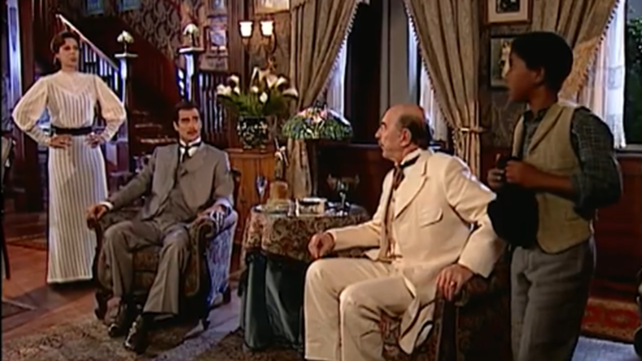 Cena mostra quatro pessoas em uma sala de estar decorada em estilo antigo. Um homem de terno claro está sentado e olha para um menino em pé, que segura um chapéu nas mãos. Outro homem, de terno cinza, também está sentado, enquanto uma mulher de vestido longo e claro observa de pé, com as mãos na cintura. O ambiente é elegante, com cortinas pesadas, flores e móveis de madeira escura.