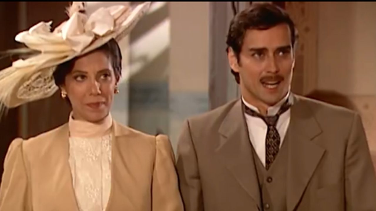 Homem e mulher vestidos com roupas de época, aparentando o início do século XX, estão lado a lado em um ambiente interno. A mulher usa um tailleur claro com gola alta rendada e um chapéu ornamentado com plumas; o homem veste terno bege, camisa branca e gravata, exibindo expressão falante enquanto a acompanha.