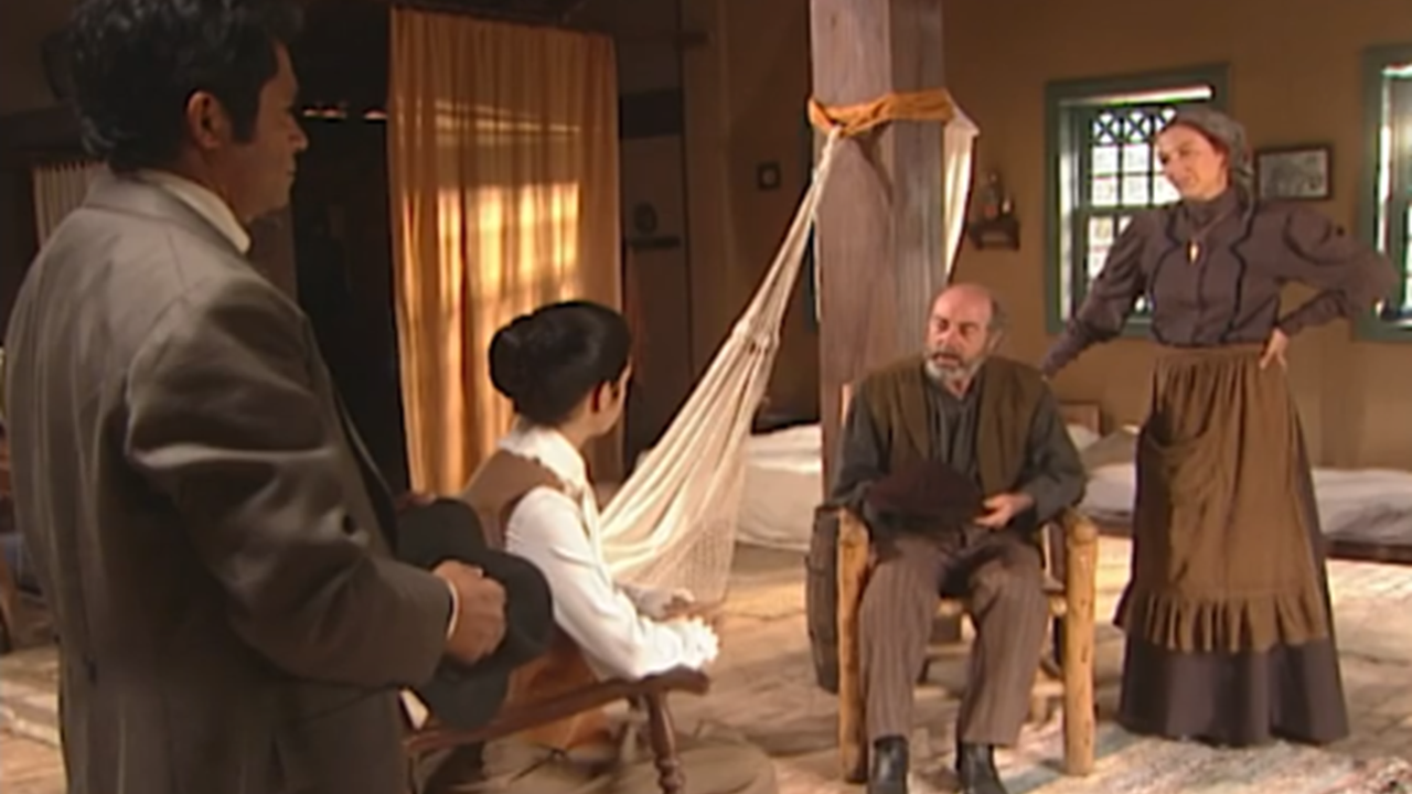 A imagem mostra uma cena ambientada em uma casa simples com decoração rústica. Quatro pessoas estão reunidas: um homem de terno segura um chapéu, uma mulher sentada o observa, enquanto um homem idoso sentado em uma cadeira parece falar, e uma mulher de roupa antiga está de pé ao lado dele, com as mãos na cintura