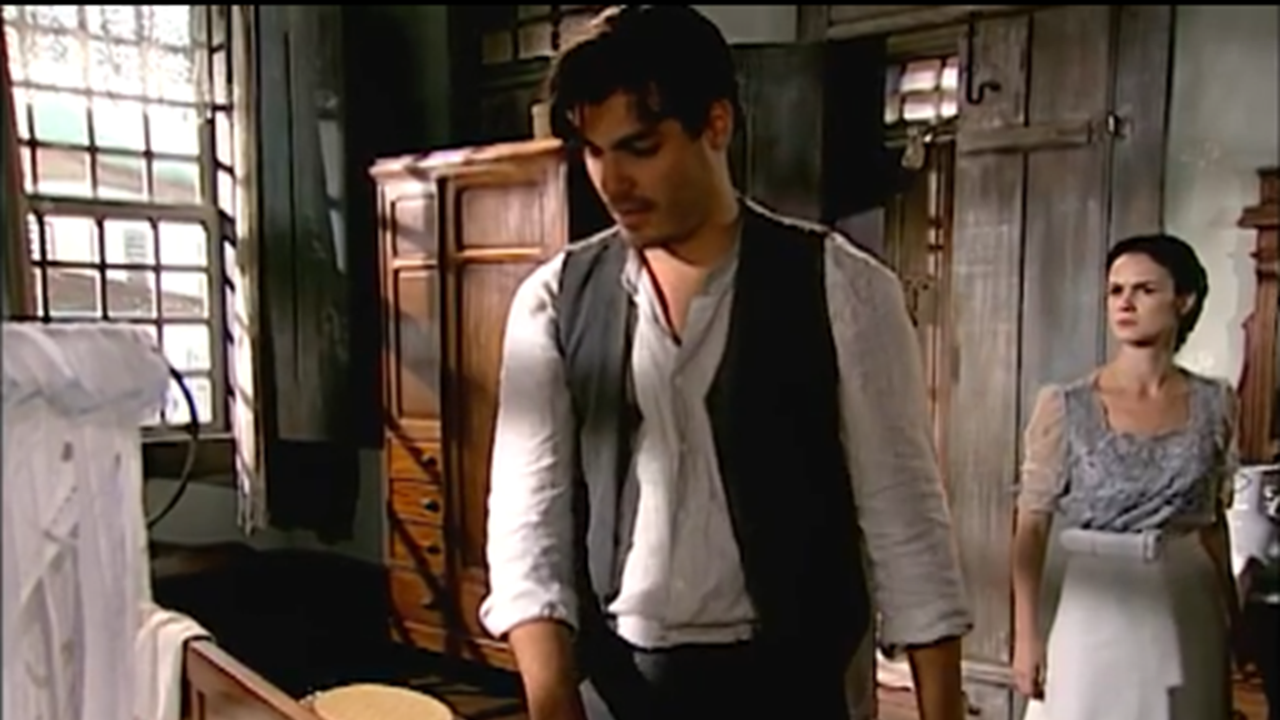 Cena de época mostra um homem e uma mulher em um quarto simples, iluminado pela luz natural que entra pelas janelas de madeira. O homem está cabisbaixo, vestindo camisa clara e colete escuro, enquanto a mulher, com expressão séria, observa ao fundo, usando um vestido antigo com avental branco