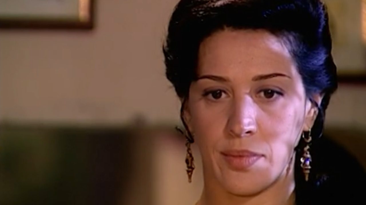 Mulher adulta aparece em close, com expressão séria e contemplativa, olhando levemente para a frente. Ela tem cabelos escuros presos para trás, maquiagem discreta e usa brincos longos e delicados. O ambiente é interno, com iluminação suave e fundo desfocado, sugerindo uma cena íntima ou dramática, possivelmente de uma produção audiovisual.