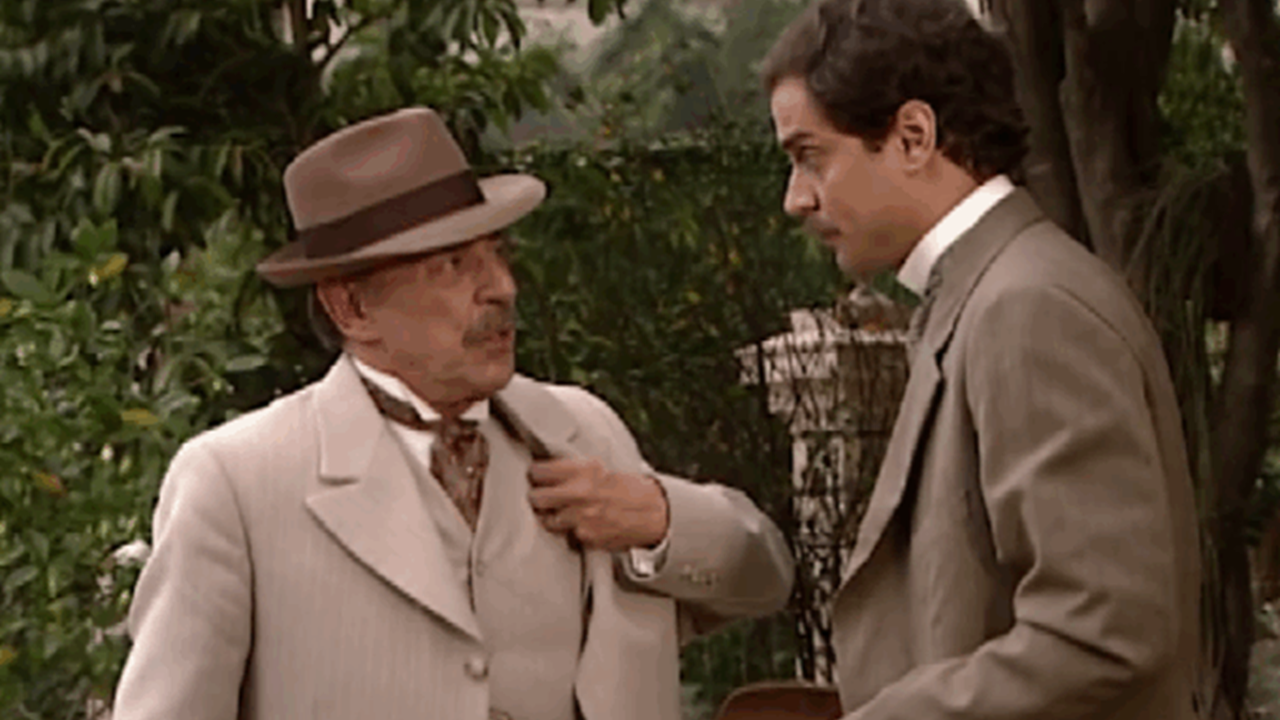 Dois homens vestidos com ternos de época conversam em um ambiente externo com vegetação ao fundo. Um deles, mais velho e usando chapéu, gesticula enquanto fala; o outro, mais jovem, escuta atentamente. A cena transmite um ar histórico e formal, típico de produções de época