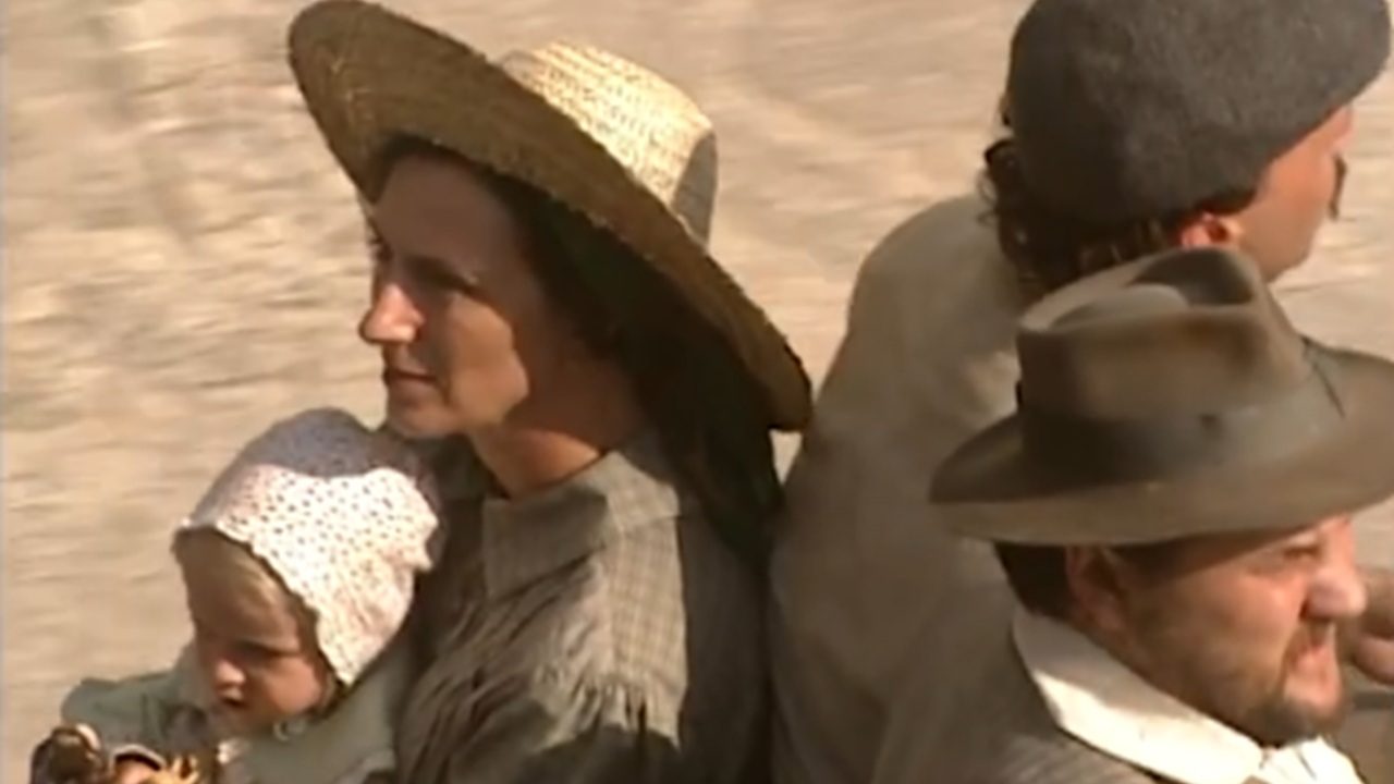 Cena ao ar livre mostra três adultos sentados de costas uns para os outros sobre uma superfície clara, em ambiente ensolarado. Em primeiro plano, uma mulher usa chapéu de palha e segura um bebê no colo, que veste touca clara. À direita, um homem usa chapéu e camisa clara, olhando para o lado oposto. Ao fundo, outro homem aparece de perfil, com gorro escuro. As roupas e o cenário sugerem um contexto rural ou de época.