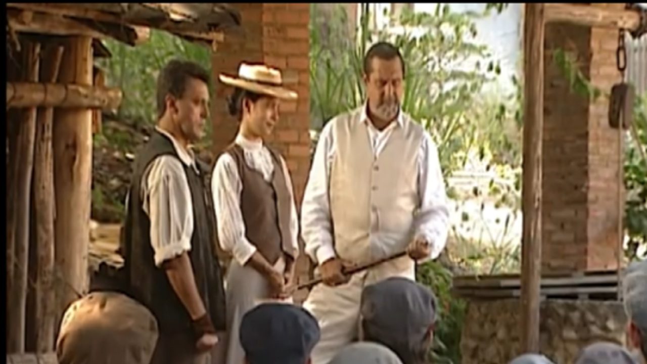 Homem de colete claro e camisa branca segura um bastão enquanto fala diante de um grupo de pessoas sentadas, acompanhado por uma mulher de chapéu e colete marrom e outro homem de colete escuro, em ambiente rústico com pilares de madeira e tijolos aparentes