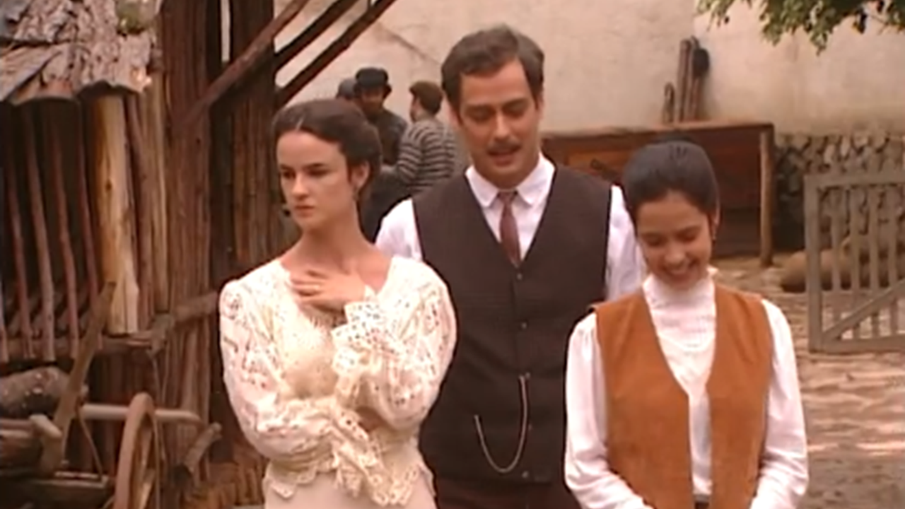 A imagem mostra três pessoas em um ambiente rural, com construções de madeira e uma carroça ao fundo. As roupas dos personagens remetem ao início do século XX: o homem veste colete e gravata, enquanto as mulheres usam vestidos e blusas de renda, com penteados presos. A cena parece fazer parte de uma produção de época, transmitindo um clima de drama e romance histórico