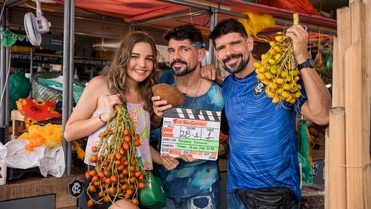 Elenco segura frutos em feira durante gravação com claquete de novela ao centro em cena de bastidores