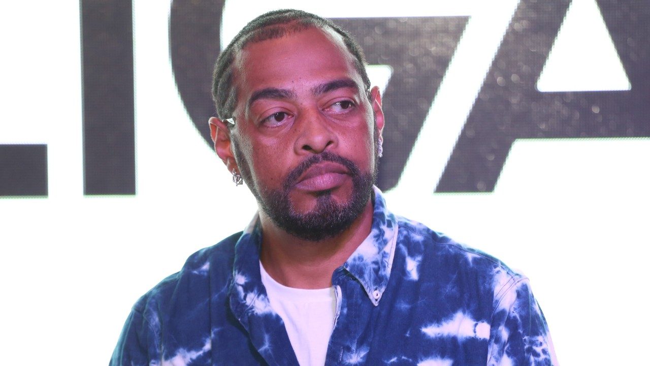 Thaíde com barba e cabelo curto olha para o lado usando camisa azul estampada diante de painel iluminado com letras ao fundo